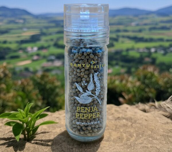 Clear glass jar of Penja Pepper with a scenic aerial view of Penja Valley farmland and mountains in the background, showcasing the premium quality of Bantu Tastes' Penja Pepper.